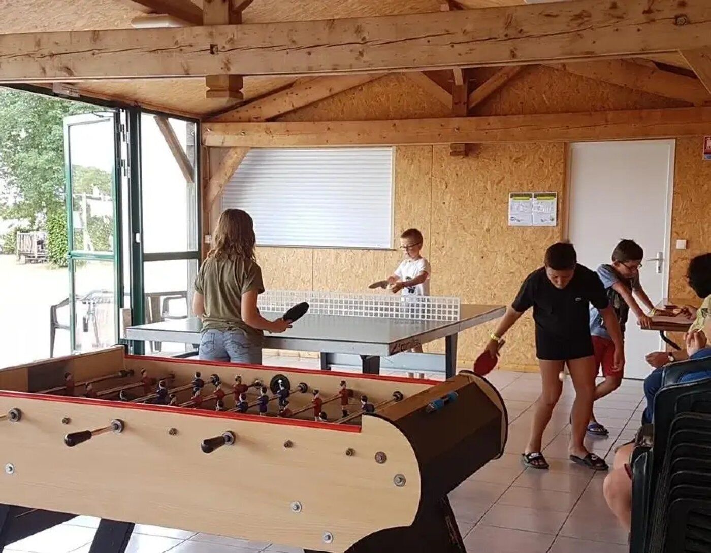 Quarto com piso de madeira, mesa de futebol de botão e mesa de pingue-pongue, e uma janela com persianas.