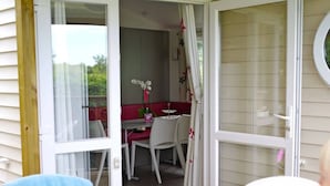 Een open glazen schuifdeur die leidt naar een eetruimte met een tafel gedekt voor twee personen, een stoel met een blauwe handdoek, en een raam met gordijnen.