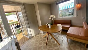 A room with a round wooden table, white chairs, a pink sofa, and a window with a view of a building. - ZEcamping 4* locatif TV LV BBQ 30m2 (Agde)
