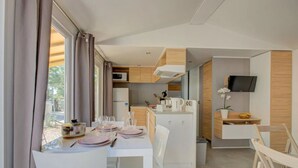 Una kitchenette compacta con microondas, un lavamanos y una pequeña encimera. Hay un comedor con sillas blancas y una mesa preparada para dos personas.