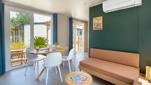 A modern living room with a sofa, a dining table with chairs, and a small round table with a magazine. - ZEcamping 4* animé 1km plage piscine chauffée locatif TV LV BBQ 31m2 (Agde)