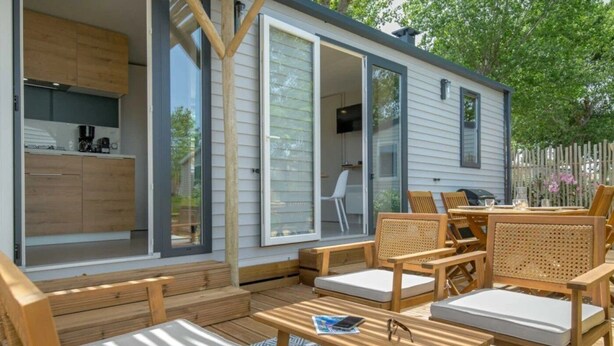 A modern outdoor living space with wooden furniture, a patio table, and a sliding glass door leading to a kitchen area. - ZEcamping 4* animé 1km plage piscine chauffée locatif TV LV BBQ 31m2 (Agde)