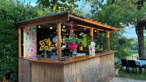 Un bar extérieur de style rustique, doté d’un comptoir en bois, entouré de verdure et d’arbres.