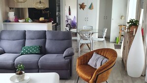 A modern living room with a grey sofa, a brown armchair, and a green armchair. There is a dining table with chairs, a kitchen area, and a white cabinet.