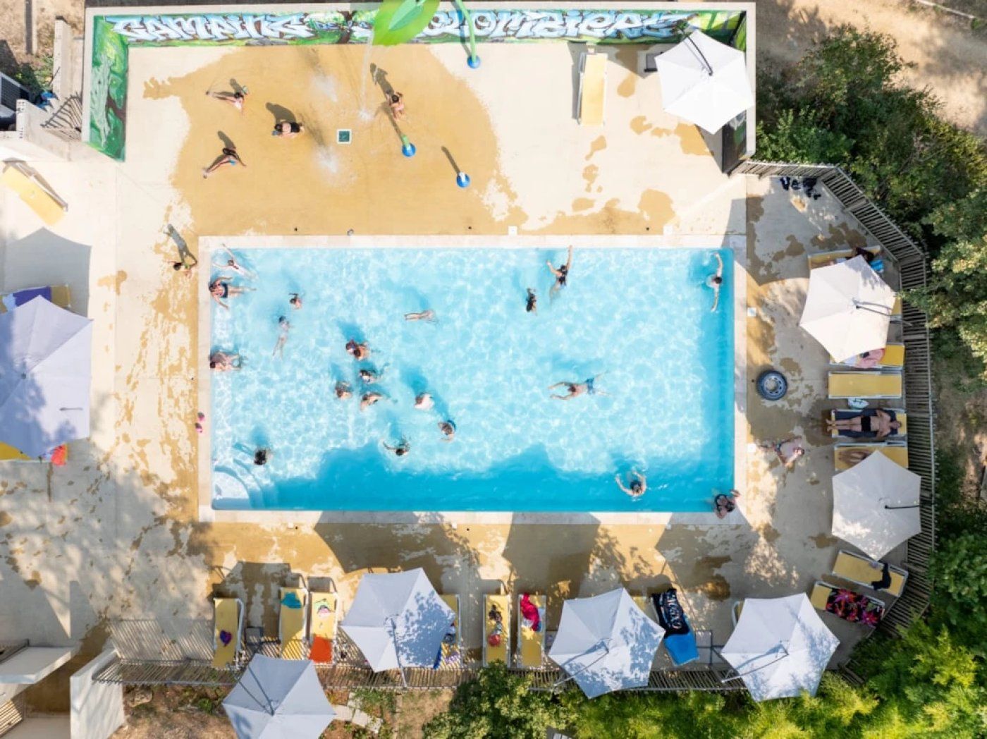 En vy från ovan av en swimmingpool där folk badar, omgiven av solstolar och parasoller.