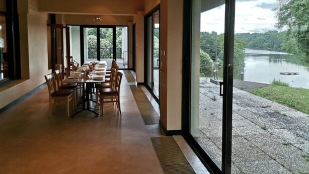 Un espace repas avec une longue table et des chaises, de grandes fenêtres donnant sur un lac et la verdure environnante.