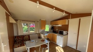 A compact kitchen with a dining area, a window with curtains, and a view of greenery outside. - ZEcamping 2 étoiles piscine chauffée mobil-home TV 25m2 5 pers (Prailles-La Couarde)