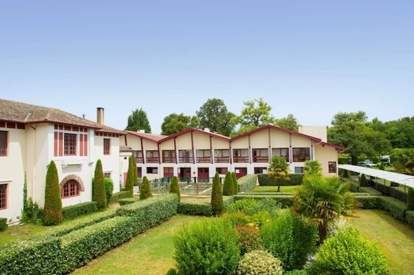 Ein Hotel mit einer symmetrischen Fassade, zahlreichen Fenstern und einem gepflegten Garten mit Hecken und Bäumen.