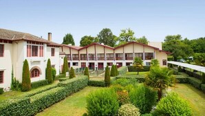 Ein Hotel mit einer symmetrischen Fassade, zahlreichen Fenstern und einem gepflegten Garten mit Hecken und Bäumen.