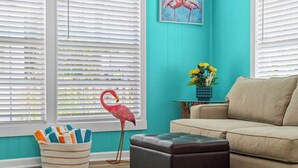 Un salon aux murs de couleur turquoise, avec un canapé beige, un pouf marron, une statue de flamant rose, un panier à rayures et un vase rempli de fleurs.