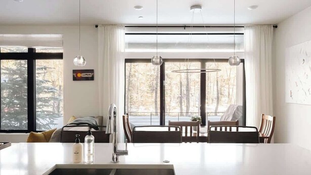 Une cuisine moderne dotée d’un îlot central, de luminaires suspendus et de grandes fenêtres offrant une vue sur les arbres.