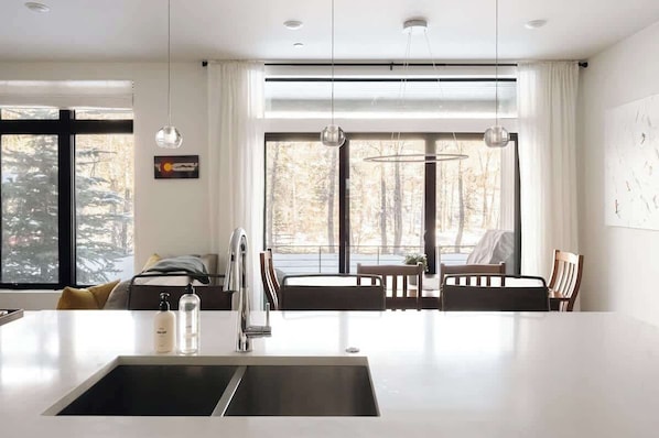 A modern kitchen with a central island, pendant lighting, and large windows with a view of trees.