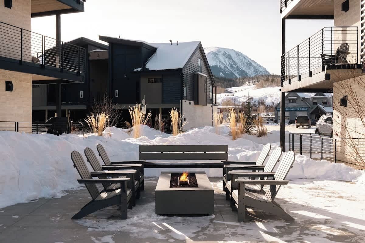 A modern outdoor seating area with Adirondack chairs and a fire pit, surrounded by snow and buildings.