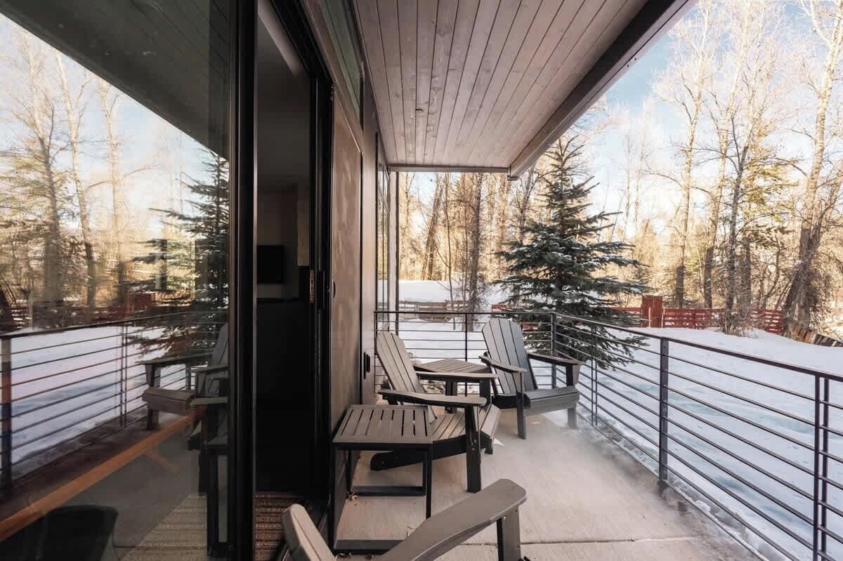 A balcony with outdoor seating, a view of a snowy forest, and a metal railing.
