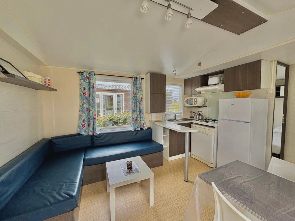 En kompakt opholdsstue med sofa, lille spisebord, tekøkken og et vindue med gardiner.