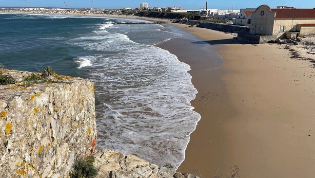A coastal beach with waves, a rocky cliff, and a building with a red roof.