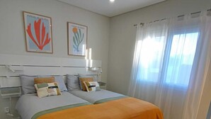 A bedroom with two beds, a headboard, a window with curtains, and two framed pictures on the wall.
