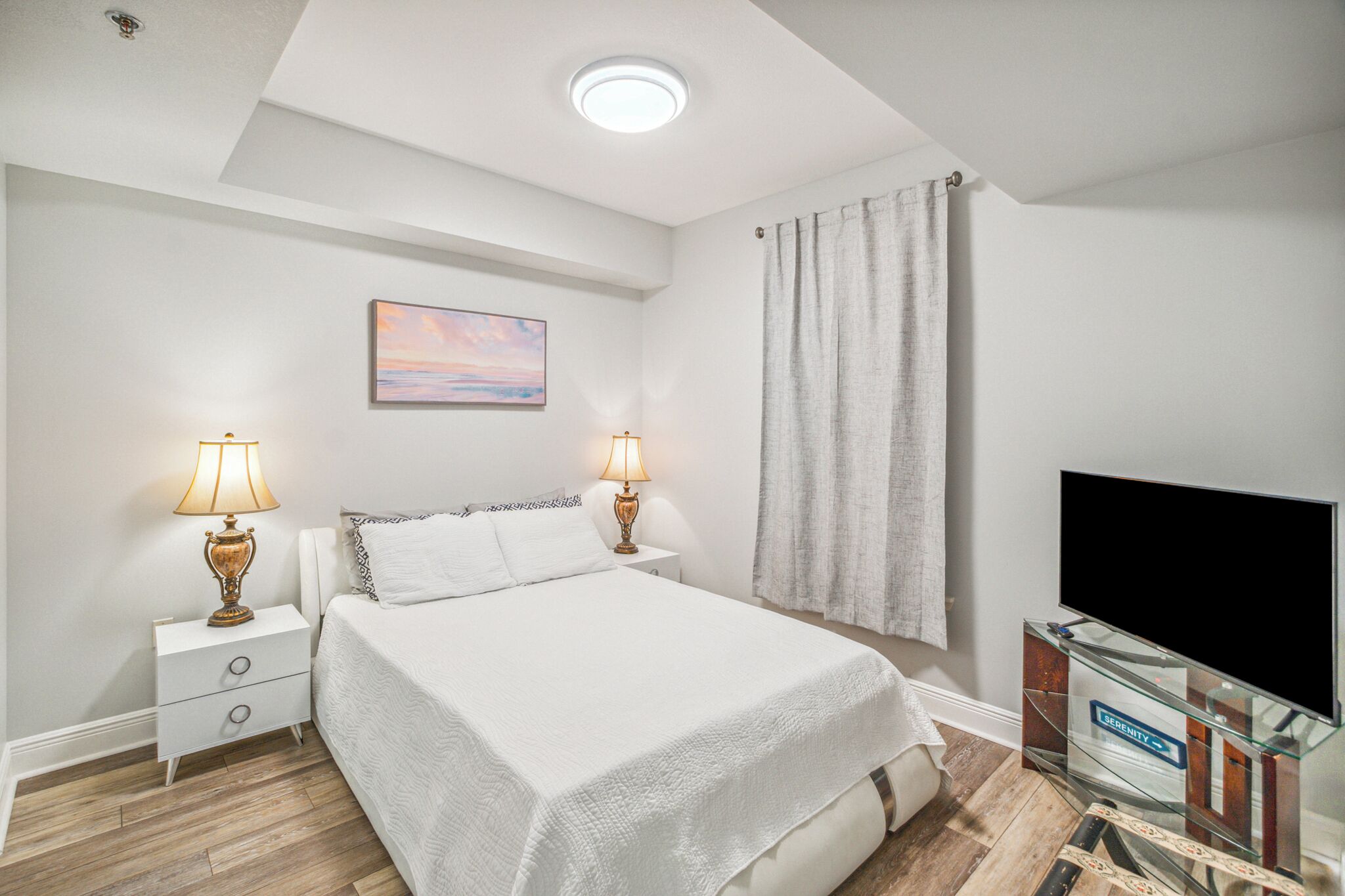 A bedroom with a bed, two bedside lamps, a painting, a television, and a curtain.