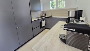 Une cuisine moderne avec des meubles gris, un four encastré, une machine à café et une fenêtre donnant sur un paysage verdoyant.
