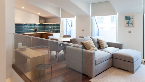 A modern interior with a living area, kitchen, and a glass railing overlooking a wooden staircase. - FLH Glória 70  Duplex I (Lisbon)