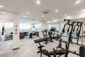 Une salle de sport moderne équipée de divers appareils d’exercice, avec un espace détente et une cuisine visible au fond.