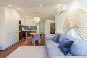 Ein modernes Wohnzimmer mit grauem Sofa, einem Esstisch mit Stühlen und einer Küchenzeile mit blauen Fliesen und Holzschränken.