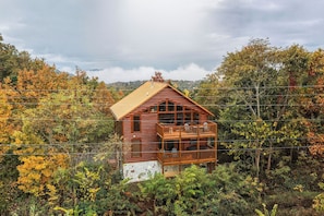 A log cabin with a balcony, surrounded by dense forest and autumn foliage.