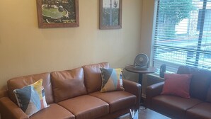 A living room with a brown leather sofa, two framed pictures on the wall, a small table with a fan, and a window with blinds.