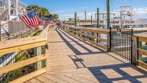 A wooden boardwalk with American flags, leading to a marina with boats and a building with a balcony. - Fall and early winter weeks available at Tupelo Bay 3BR Villa! (Myrtle Beach)