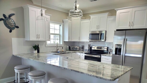 A modern kitchen with white cabinets, a large island with stools, stainless steel appliances, and a decorative turtle wall art piece. - Fall and early winter weeks available at Tupelo Bay 3BR Villa! (Myrtle Beach)
