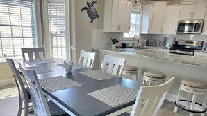 A modern kitchen with white cabinetry, a central island, and a dining area with white chairs and placemats. - Fall and early winter weeks available at Tupelo Bay 3BR Villa! (Myrtle Beach)