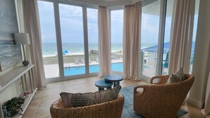A room with a large window overlooking a beach and pool area. There are wicker chairs and a small round table.