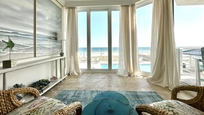 A room with a large window overlooking a beach, wicker furniture, and a painting of the ocean.