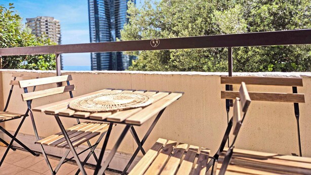 A balcony with a table and chairs, overlooking a cityscape with tall buildings and greenery. - LX146 - Studio cosy avec balcon & vue mer - Monaco (Beausoleil)
