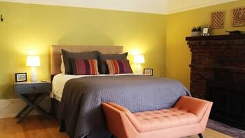 A bedroom with a bed, two bedside tables with lamps, a brick fireplace, and a pink armchair.