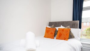 Una cama bien hecha con sábanas blancas y almohadas anaranjadas estampadas, una toalla blanca enrollada y vista a un edificio de ladrillo a través de una ventana con cortinas azules.