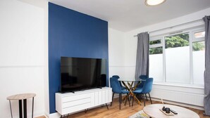Una sala de estar moderna con un televisor de pantalla plana grande, un armario blanco, dos sillas azules, una mesita redonda y una ventana con cortinas.