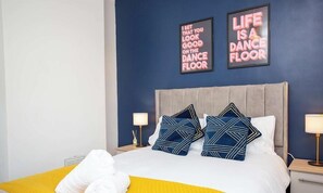 A hotel room with a bed, two wall art pieces, a nightstand with a lamp, and a yellow bedspread.