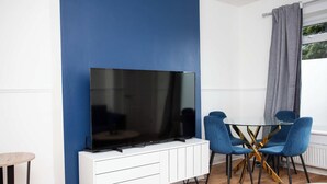Una habitación con una televisión de pantalla plana grande sobre un mueble blanco, dos sillas azules alrededor de una mesa de vidrio y una ventana con cortinas grises.
