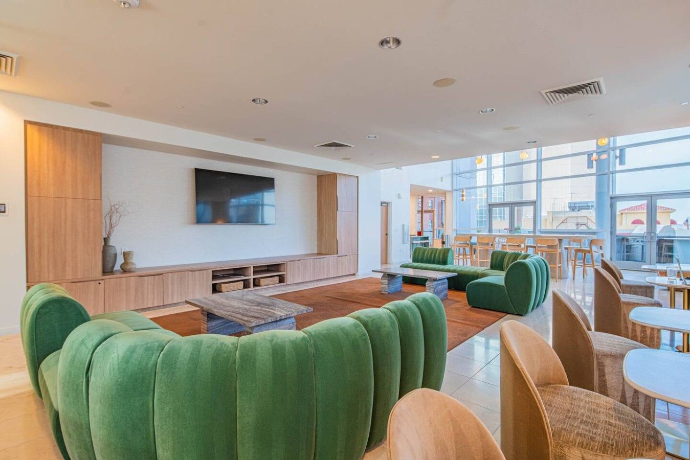 A spacious lounge area with green upholstered seating, a flat-screen TV mounted on the wall, and a large window offering a view of the city.