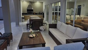 Un salon moderne avec un canapé, des fauteuils et une table basse. On aperçoit à l’arrière-plan un coin cuisine avec une table à manger et des chaises.
