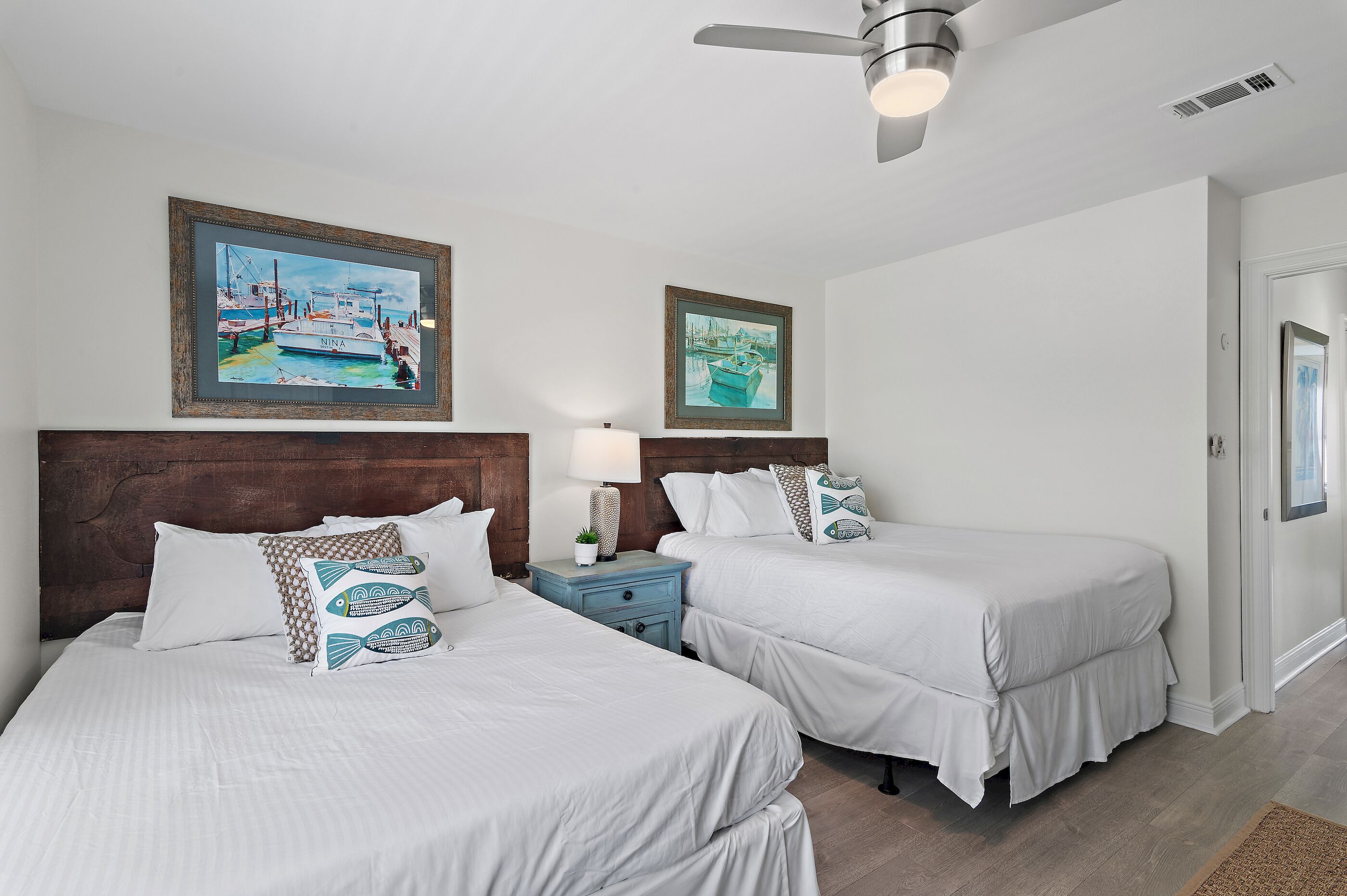 A hotel room with two beds, wooden headboards, white bedding, a nightstand with a lamp, and two framed paintings on the wall.