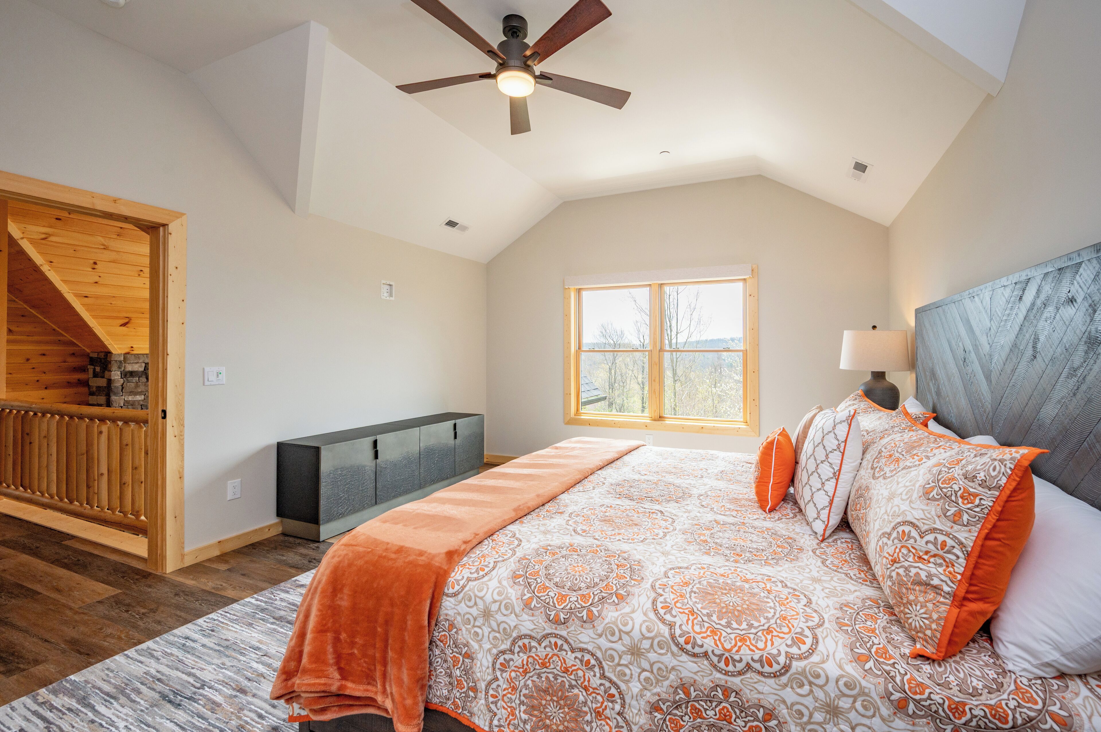 A bedroom with a large bed, a ceiling fan, a window, and a wooden staircase.