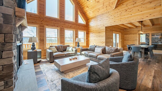 A spacious log cabin interior with a large living area, stone fireplace, and multiple windows. - Spacious Lodge w/ Hot Tub, Fire Pit & Games (Swanton)