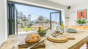 A kitchen with a large window, a wooden table with a loaf of bread, a bowl of salad, a pizza on a wooden board, and a bottle of wine. - One Beach Road - sleeps 10 guests  in 5 bedrooms (Rhosneigr)