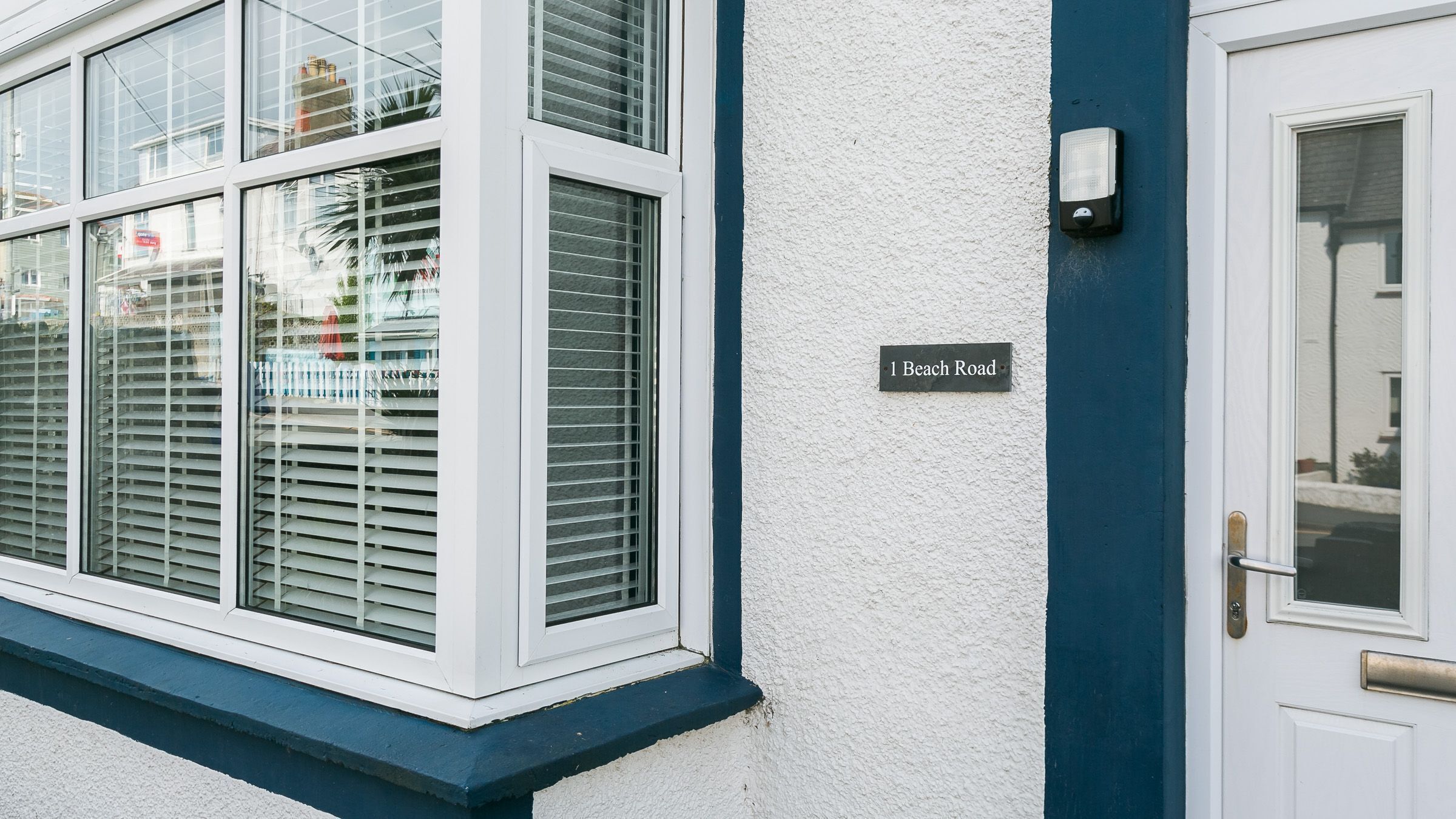 A house with a white exterior, a blue trim around the windows, and a door with a brass handle. A sign on the wall reads "1 Beach Road."