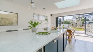 A modern kitchen with a large island, a dining area with a wooden table, and a view of the garden through large windows. - One Beach Road - sleeps 10 guests  in 5 bedrooms (Rhosneigr)
