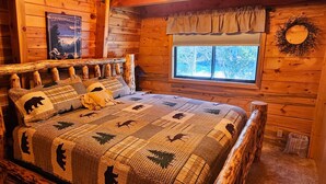 Ein gemütliches Schlafzimmer in einer Blockhütte mit einem Bett, das eine Bettdecke mit Waldmuster hat, einem Fenster mit Blick auf Bäume und einem an der Wand befestigten Schild mit der Aufschrift "HELTON VALLEY".