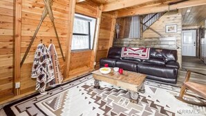 Ein Holzblockhaus-Interieur mit einem Ledersofa, einem Esstisch mit Teller und Tassen, einem gemusterten Teppich und einer Treppe, die zu einer weiteren Ebene führt.