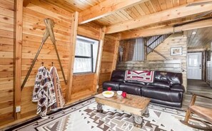 L’intérieur d’une cabane en rondins, avec un canapé en cuir, une table à manger avec une assiette et des tasses, un tapis à motifs et un escalier menant à un autre niveau.